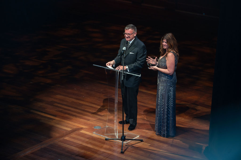 Two presenters at the podium on stage, woman holding trophy