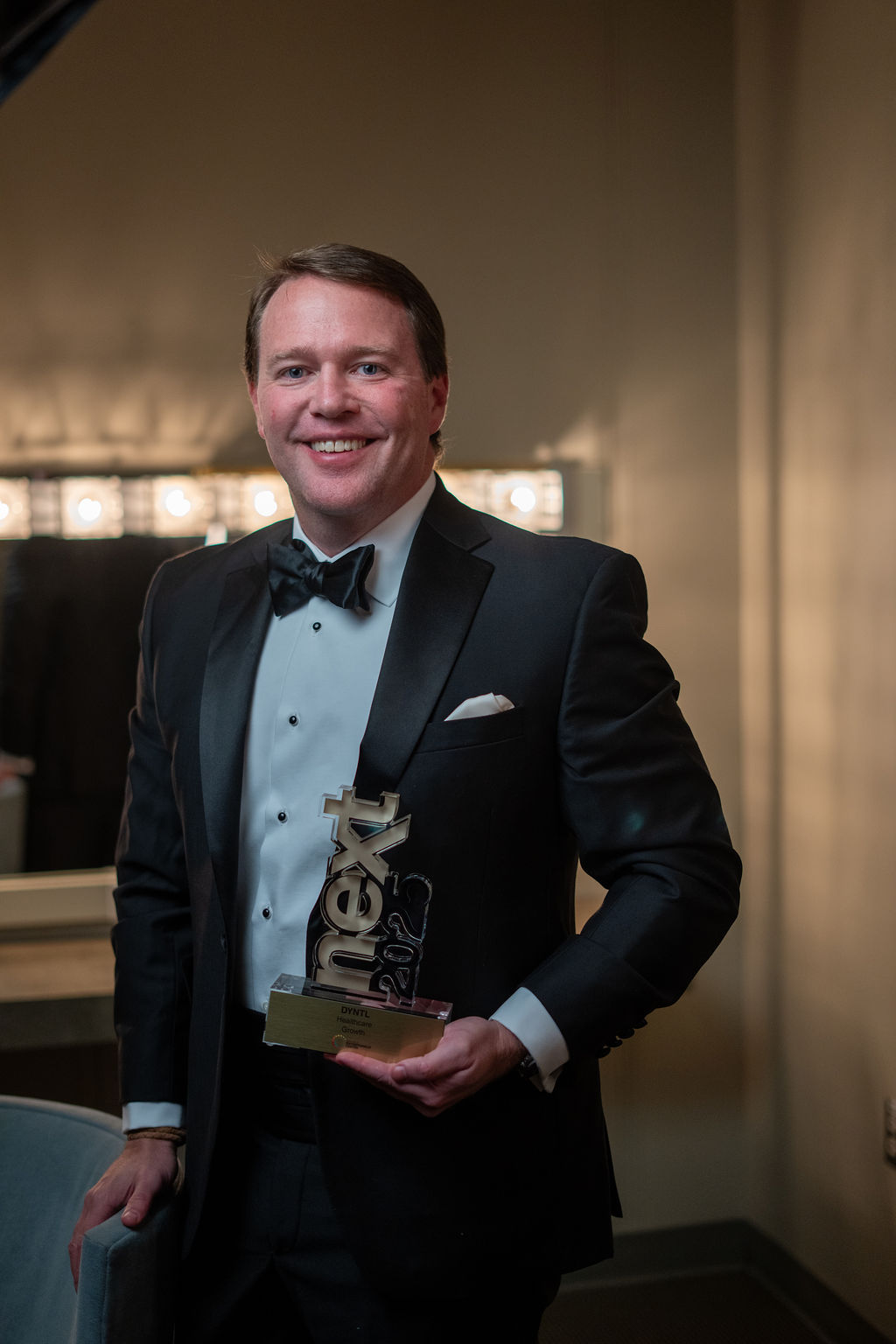 Man in tuxedo with NEXT trophy, confident portrait in dressing room