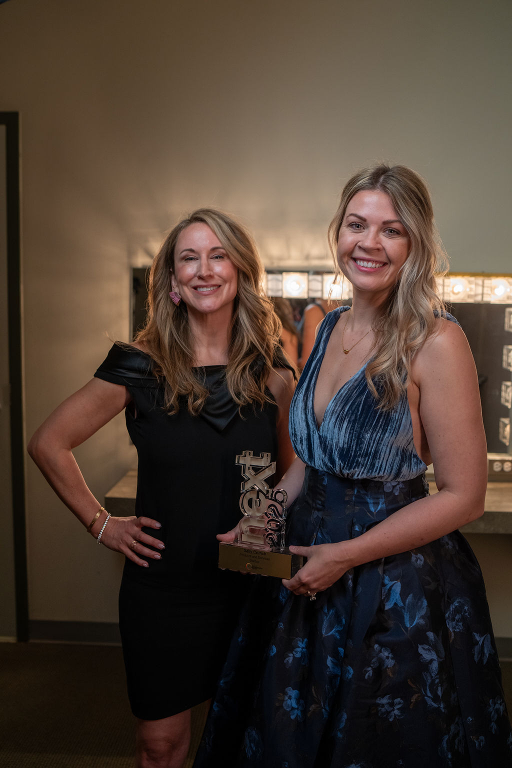 Two women smiling with NEXT trophy in front of dressing room mirror lights