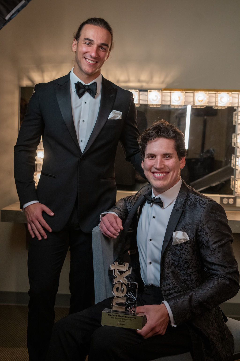 Two men in tuxedos smiling with NEXT trophy in dressing room