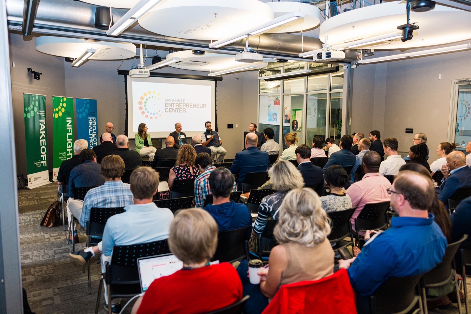 A full InFlight cohort session at the EC, founders seated at tables facing speakers at the front of the room, InFlight and TakeOff banners beside the screen