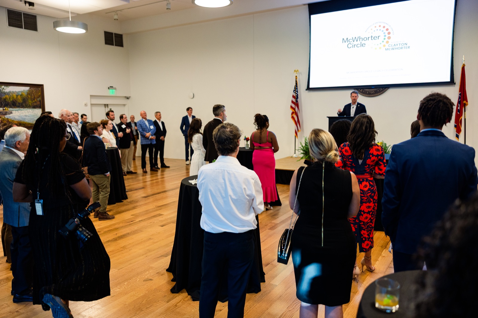 Guests standing in a bright hall facing a speaker at a podium beneath the McWhorter Circle mark, with the U.S. and Tennessee flags beside the screen
