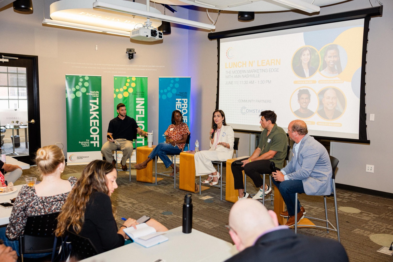 Members seated at tables watching a five-person panel in front of program banners
