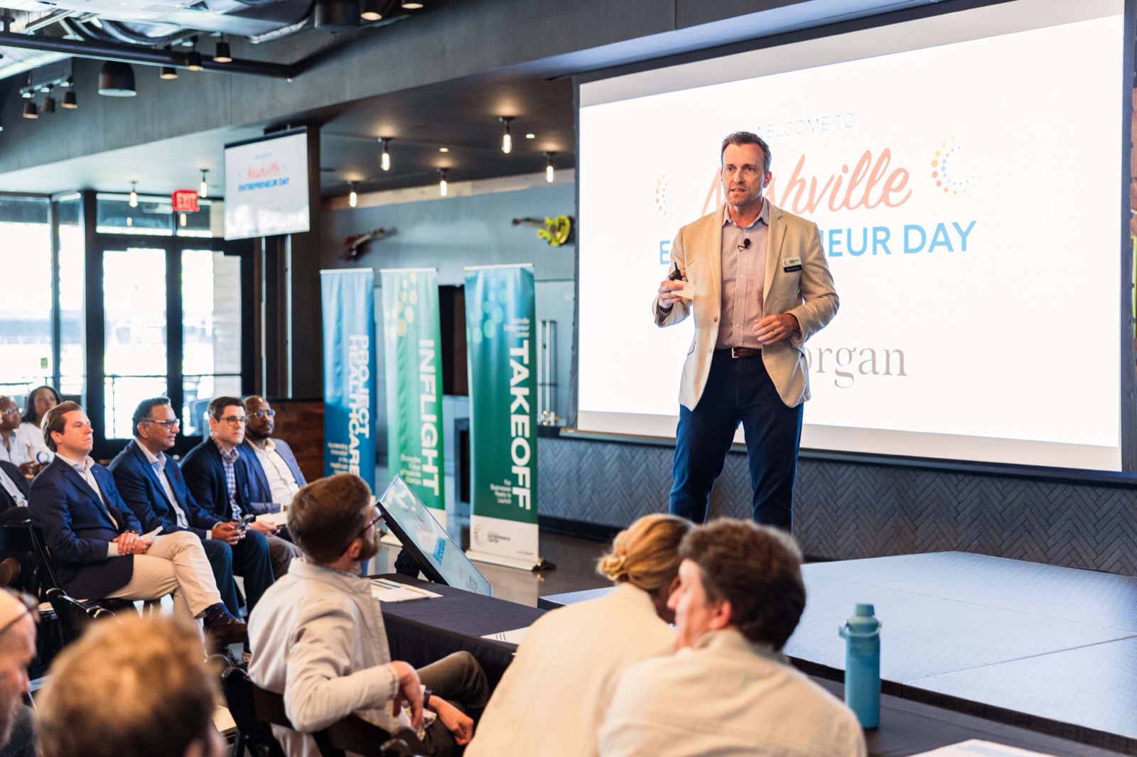 A speaker on stage in front of a Welcome to Nashville Entrepreneur Day slide, with TakeOff, InFlight, and Project Healthcare banners beside the stage