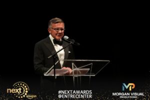 A man in a tuxedo and glasses speaks at a clear podium on stage at the 2026 NEXT Awards.