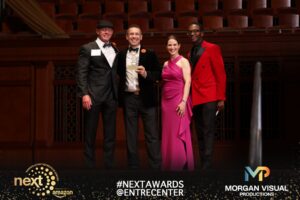 A winner holds a NEXT 2026 trophy on stage, smiling with three people in formal wear.