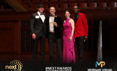 A winner holds a NEXT 2026 trophy on stage, smiling with three people in formal wear.