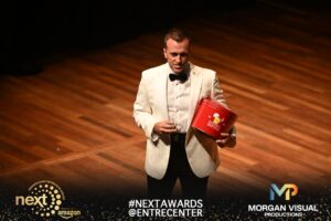 A man in a white tuxedo jacket holds a red Kernels Nashville Popcorn tin on stage.