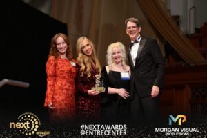 Three women and a man in a tuxedo pose on stage, one woman holding a NEXT trophy.