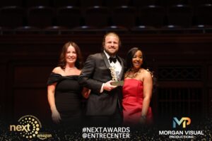 A man in a tuxedo holds a NEXT 2026 trophy on stage between two women.